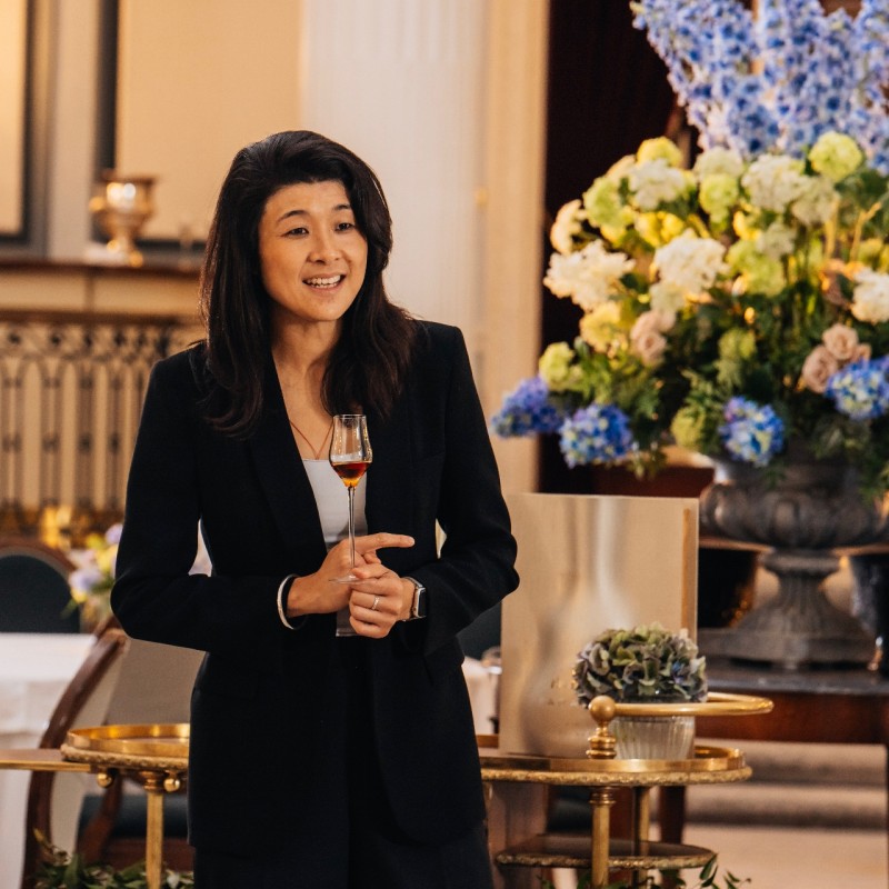 Woman in formal attire holding a glass of wine in an elegant indoor setting with floral arrangements.